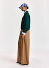 A model shown in profile wearing a dark green knit cardigan with black sequined embroidery, a light blue collared shirt underneath, wide-leg tan trousers, and a blue and white plaid baseball cap, set against a plain light gray studio background.