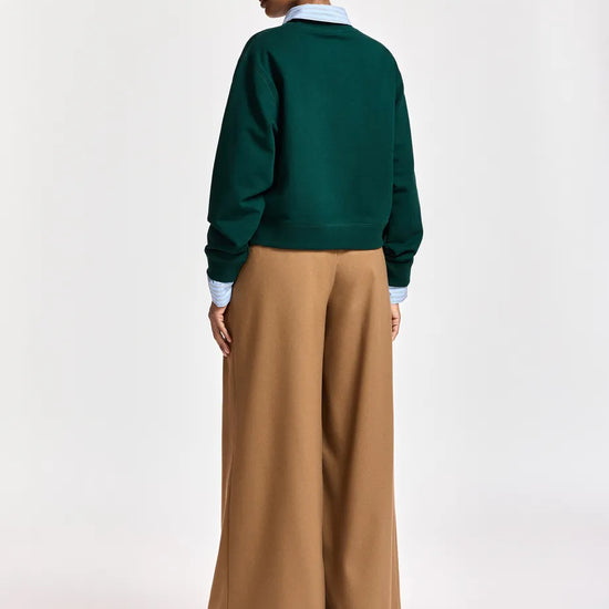 A model shown from the back wearing a dark green crewneck sweater, wide-leg tan trousers, a light blue collared shirt, a blue and white plaid cap, and black shoes, against a plain light gray studio background.