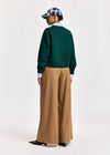 A model shown from the back wearing a dark green crewneck sweater, wide-leg tan trousers, a light blue collared shirt, a blue and white plaid cap, and black shoes, against a plain light gray studio background.