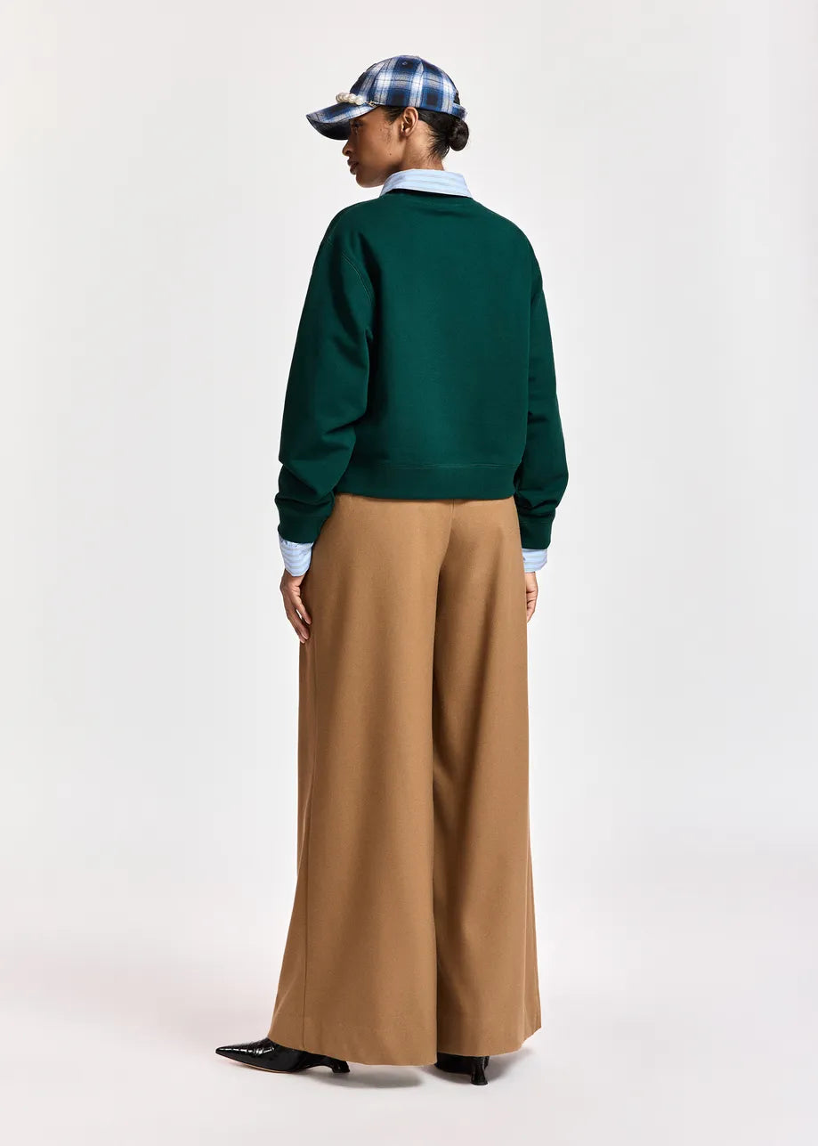A model shown from the back wearing a dark green crewneck sweater, wide-leg tan trousers, a light blue collared shirt, a blue and white plaid cap, and black shoes, against a plain light gray studio background.