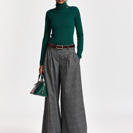 A model stands against a light gray studio background wearing a dark green ribbed turtleneck sweater, high-waisted wide-leg gray trousers with a brown belt, dark sunglasses, and holding a small green handbag.