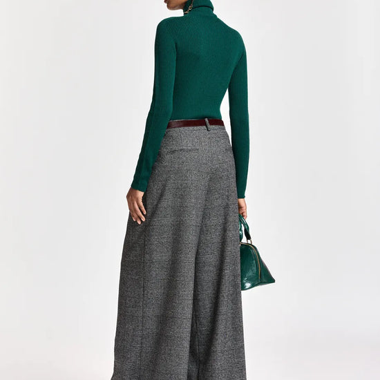 A model is shown from a rear three-quarter view wearing a dark green long-sleeve mock neck top, high-waisted gray wide-leg pleated trousers, a dark brown belt, and burgundy pointed-toe flats. She is holding a small green handbag against a plain white studio background.