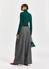 A model is shown from a rear three-quarter view wearing a dark green long-sleeve mock neck top, high-waisted gray wide-leg pleated trousers, a dark brown belt, and burgundy pointed-toe flats. She is holding a small green handbag against a plain white studio background.