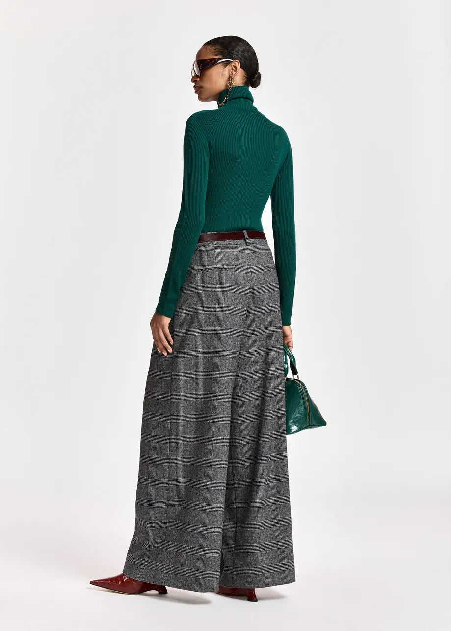 A model is shown from a rear three-quarter view wearing a dark green long-sleeve mock neck top, high-waisted gray wide-leg pleated trousers, a dark brown belt, and burgundy pointed-toe flats. She is holding a small green handbag against a plain white studio background.