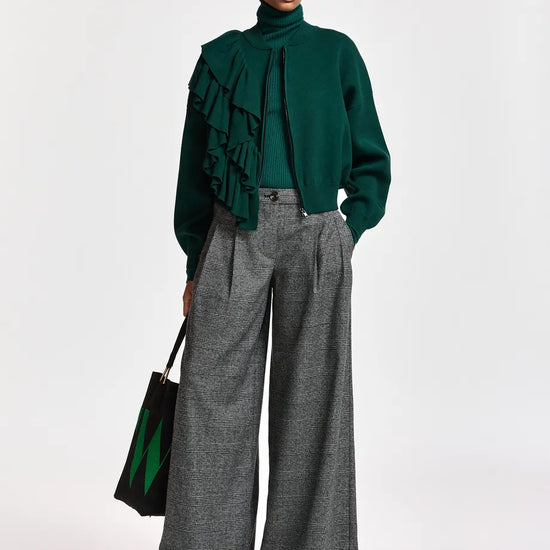 A model stands against a light gray studio background wearing a dark green ruffled turtleneck sweater, wide-leg gray trousers, and red pointed shoes, while holding a black tote bag with green accents.
