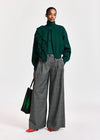 A model stands against a light gray studio background wearing a dark green ruffled turtleneck sweater, wide-leg gray trousers, and red pointed shoes, while holding a black tote bag with green accents.