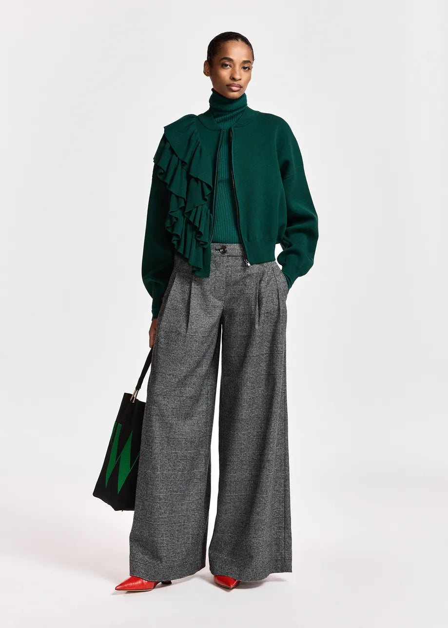 A model stands against a light gray studio background wearing a dark green ruffled turtleneck sweater, wide-leg gray trousers, and red pointed shoes, while holding a black tote bag with green accents.