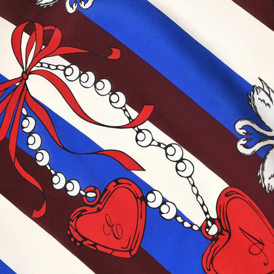 A close-up of a printed textile or scarf pattern with diagonal blue and dark red stripes, white decorative motifs, a red ribbon bow, a beaded chain, and two red heart-shaped medallions.