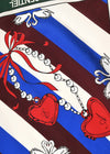 A close-up of a printed textile or scarf pattern with diagonal blue and dark red stripes, white decorative motifs, a red ribbon bow, a beaded chain, and two red heart-shaped medallions.