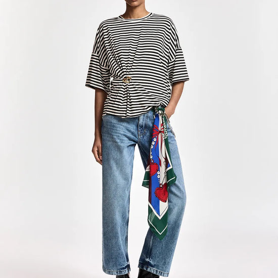A model stands against a light gray studio background wearing a black and white horizontally striped short-sleeve top, light-wash blue jeans with a colorful patterned scarf tied around the waist, and black ankle boots.