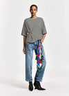 A model stands against a light gray studio background wearing a black and white horizontally striped short-sleeve top, light-wash blue jeans with a colorful patterned scarf tied around the waist, and black ankle boots.