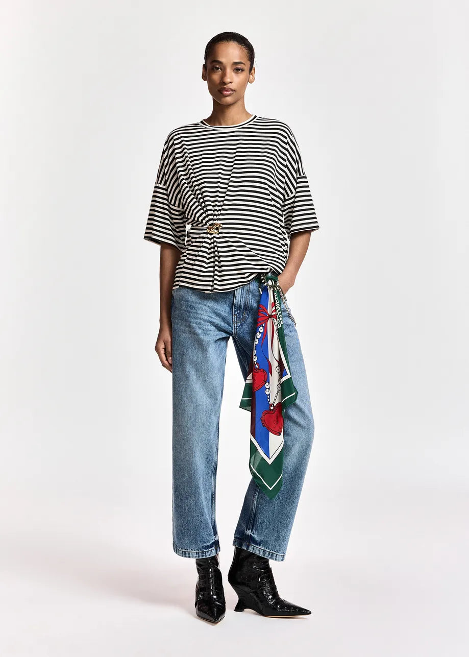 A model stands against a light gray studio background wearing a black and white horizontally striped short-sleeve top, light-wash blue jeans with a colorful patterned scarf tied around the waist, and black ankle boots.