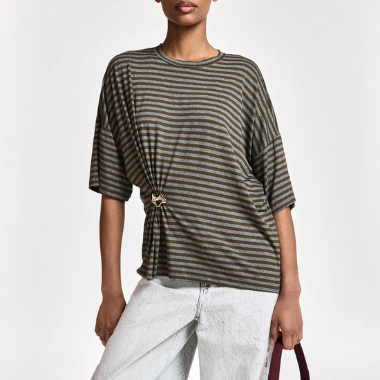 A model wearing a dark olive green and black horizontally striped short-sleeve top with a twisted front detail and a gold-tone ring clasp, paired with light-wash wide-leg jeans and holding a burgundy tote bag.