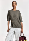 A model wearing a dark olive green and black horizontally striped short-sleeve top with a twisted front detail and a gold-tone ring clasp, paired with light-wash wide-leg jeans and holding a burgundy tote bag.