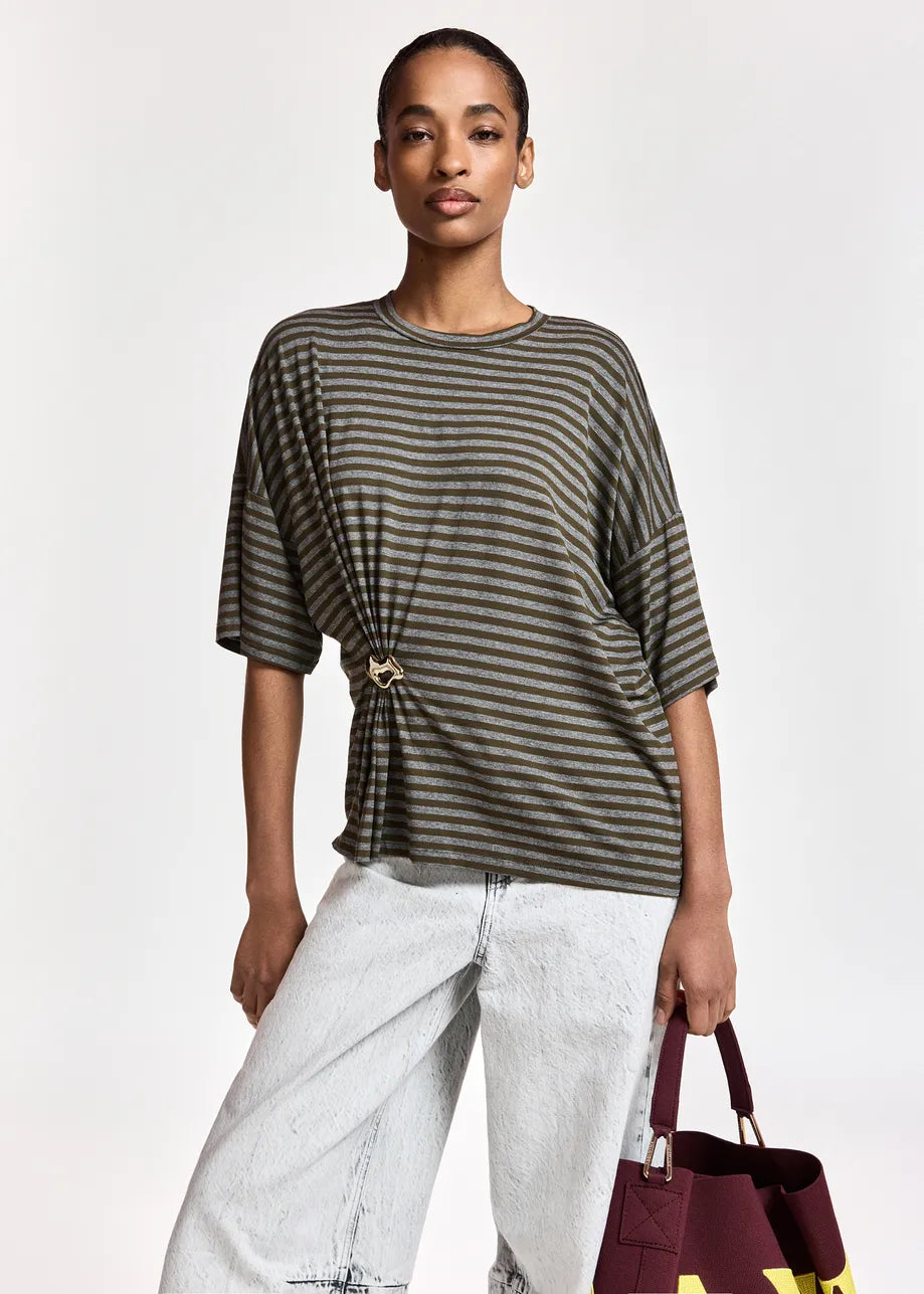 A model wearing a dark olive green and black horizontally striped short-sleeve top with a twisted front detail and a gold-tone ring clasp, paired with light-wash wide-leg jeans and holding a burgundy tote bag.
