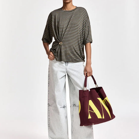 A model stands against a light gray studio background wearing a dark olive green and gray striped t-shirt, light-wash wide-leg jeans, and holding a burgundy tote bag with yellow lettering.