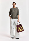 A model stands against a light gray studio background wearing a dark olive green and gray striped t-shirt, light-wash wide-leg jeans, and holding a burgundy tote bag with yellow lettering.