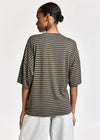 A woman shown from the back wearing an oversized short-sleeve top with horizontal olive green and dark gray stripes, paired with light gray pants, against a plain light gray studio background.