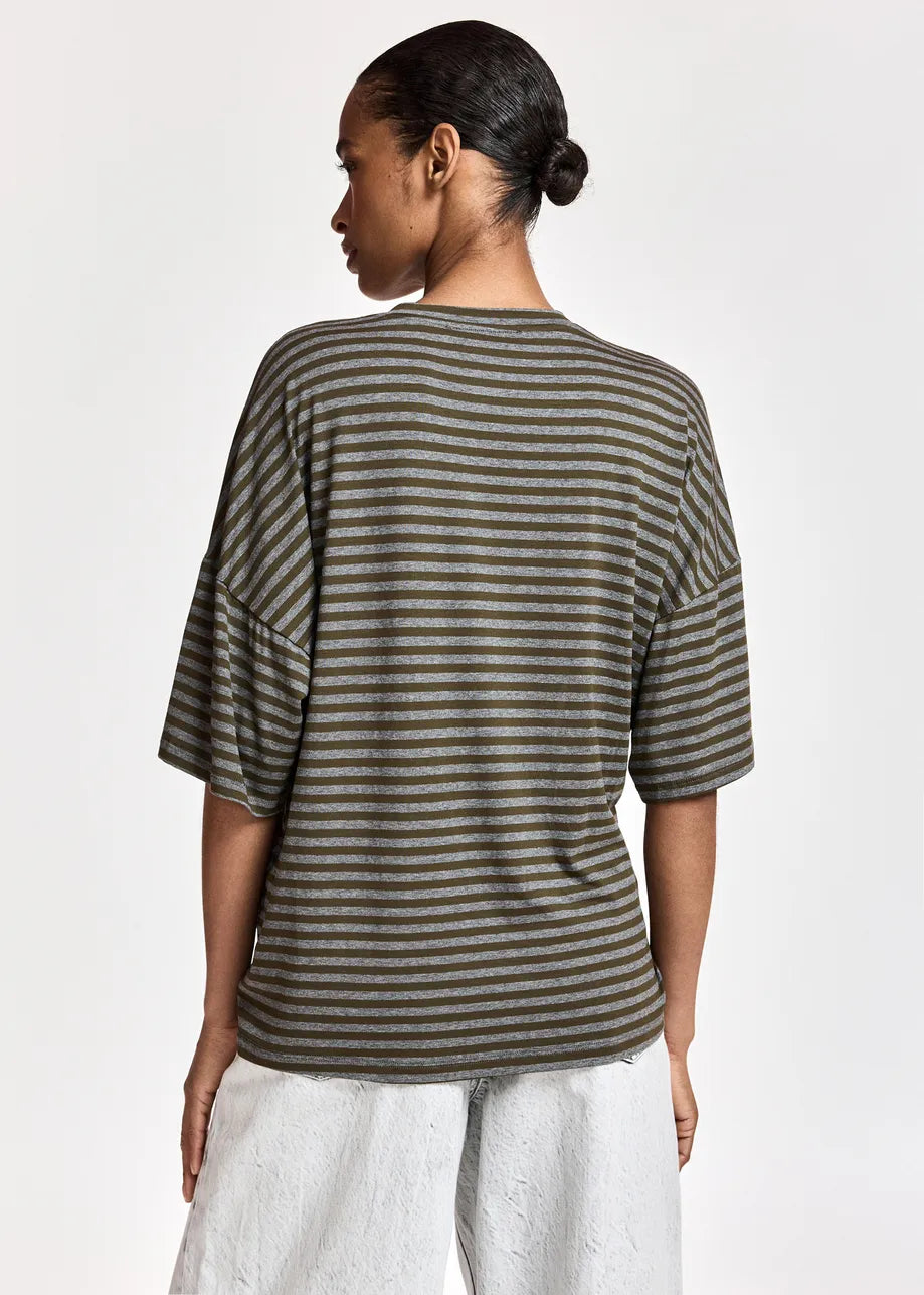 A woman shown from the back wearing an oversized short-sleeve top with horizontal olive green and dark gray stripes, paired with light gray pants, against a plain light gray studio background.