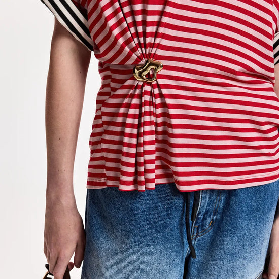 A close-up of a person wearing a red and white striped short-sleeve top with a gold-tone decorative clasp at the waist, paired with blue denim shorts, against a plain light background.