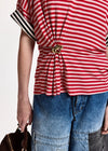 A close-up of a person wearing a red and white striped short-sleeve top with a gold-tone decorative clasp at the waist, paired with blue denim shorts, against a plain light background.