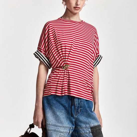 A model wearing a red and white striped short-sleeve top with black and white striped cuffs, paired with blue denim patchwork pants and holding a brown leather bag against a light gray background.