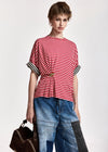 A model wearing a red and white striped short-sleeve top with black and white striped cuffs, paired with blue denim patchwork pants and holding a brown leather bag against a light gray background.