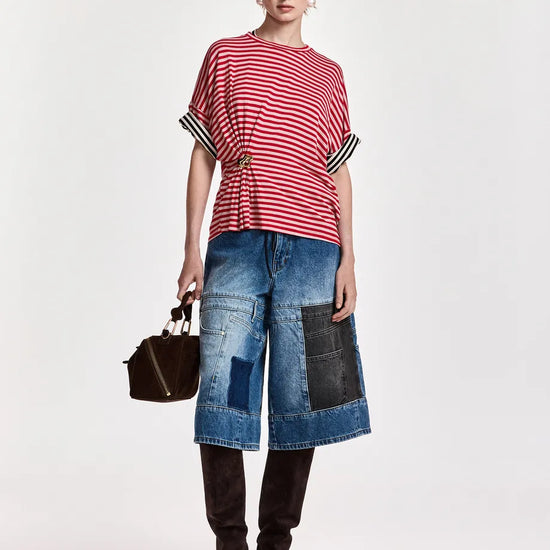 A model stands against a plain light background wearing a red and white striped short-sleeve top with black-and-white striped cuffs, blue denim patchwork cropped pants, black boots, and holding a small black handbag.