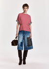 A model stands against a plain light background wearing a red and white striped short-sleeve top with black-and-white striped cuffs, blue denim patchwork cropped pants, black boots, and holding a small black handbag.