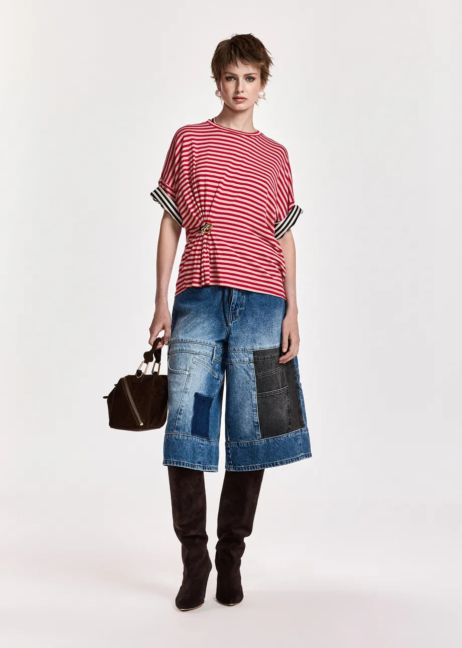 A model stands against a plain light background wearing a red and white striped short-sleeve top with black-and-white striped cuffs, blue denim patchwork cropped pants, black boots, and holding a small black handbag.