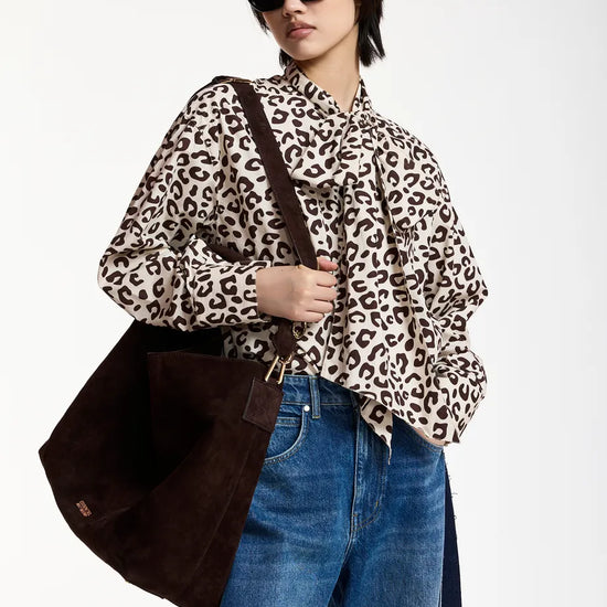 A model wearing a beige and black leopard print blouse with a ruffled collar, paired with blue jeans and a dark brown suede shoulder bag, set against a plain white studio background.
