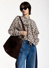 A model wearing a beige and black leopard print blouse with a ruffled collar, paired with blue jeans and a dark brown suede shoulder bag, set against a plain white studio background.