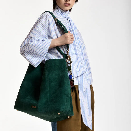 A model wearing a light blue and white striped shirt, brown trousers, and green sunglasses carries a large dark green suede tote bag with a patterned chain strap against a plain white studio background.