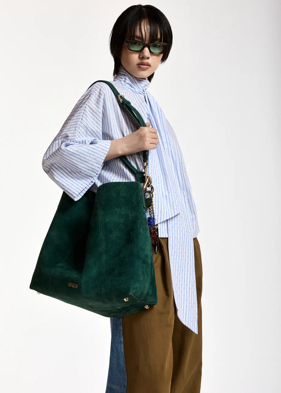 A model wearing a light blue and white striped shirt, brown trousers, and green sunglasses carries a large dark green suede tote bag with a patterned chain strap against a plain white studio background.