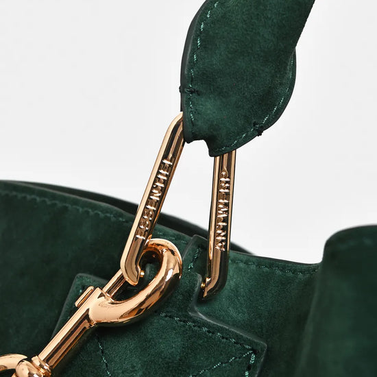 Close-up of a dark green suede handbag with gold-tone hardware and a matching green leather strap, showing engraved brand text on the clips.