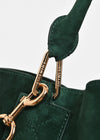 Close-up of a dark green suede handbag with gold-tone hardware and a matching green leather strap, showing engraved brand text on the clips.