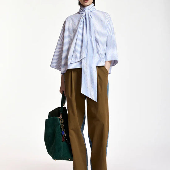 A model stands against a plain white studio background wearing a light blue and white vertically striped high-neck blouse with a large bow tie detail, brown wide-leg trousers, dark green sunglasses, and holding a dark green tote bag.
