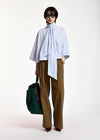 A model stands against a plain white studio background wearing a light blue and white vertically striped high-neck blouse with a large bow tie detail, brown wide-leg trousers, dark green sunglasses, and holding a dark green tote bag.