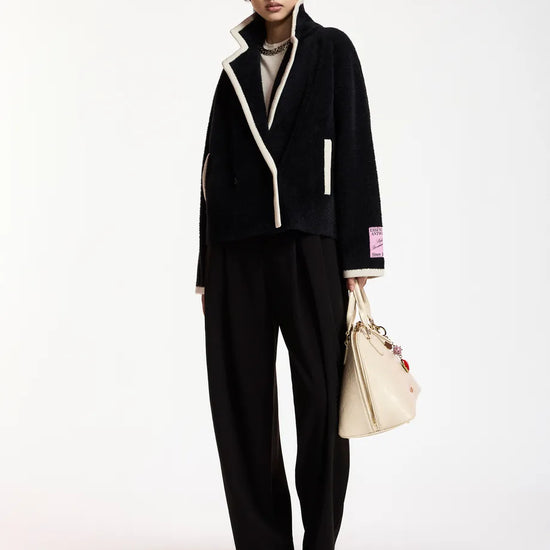A model stands against a plain white studio background wearing a black tailored jacket with contrasting cream trim, matching black wide-leg trousers, a gray top, black shoes, and carrying a cream tote bag.