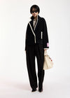 A model stands against a plain white studio background wearing a black tailored jacket with contrasting cream trim, matching black wide-leg trousers, a gray top, black shoes, and carrying a cream tote bag.