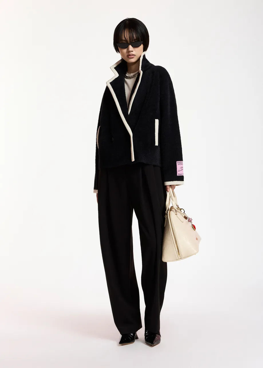 A model stands against a plain white studio background wearing a black tailored jacket with contrasting cream trim, matching black wide-leg trousers, a gray top, black shoes, and carrying a cream tote bag.