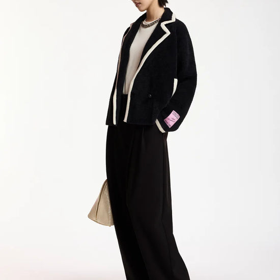 A model wearing a black velvet blazer with white piping, black wide-leg trousers, and black pointed-toe heels, standing against a plain white studio background.