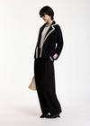 A model wearing a black velvet blazer with white piping, black wide-leg trousers, and black pointed-toe heels, standing against a plain white studio background.