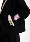 Close-up of a black fuzzy jacket with cream ribbed trim and a pink sleeve patch 