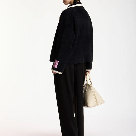 A model is shown from the back wearing a black textured jacket with white collar and cuff trim, black wide-leg trousers, and holding a beige tote bag against a plain white studio background.
