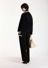 A model is shown from the back wearing a black textured jacket with white collar and cuff trim, black wide-leg trousers, and holding a beige tote bag against a plain white studio background.