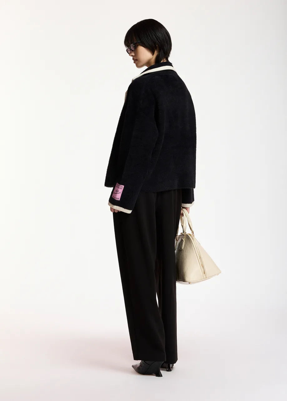 A model is shown from the back wearing a black textured jacket with white collar and cuff trim, black wide-leg trousers, and holding a beige tote bag against a plain white studio background.