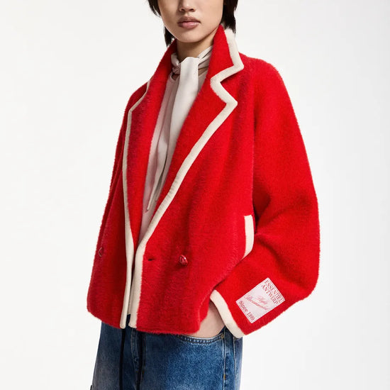 A model wearing a vibrant red fuzzy cropped jacket with cream-colored piping, paired with a white turtleneck and blue denim shorts, shot against a plain white studio background.
