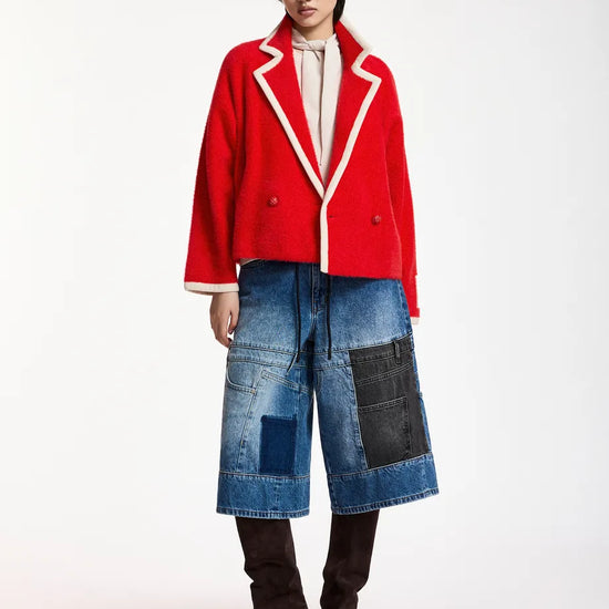 A model stands against a plain white studio background wearing a bright red cropped double-breasted blazer with white piping, blue patchwork denim culottes, and dark brown knee-high boots.