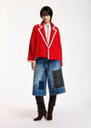 A model stands against a plain white studio background wearing a bright red cropped double-breasted blazer with white piping, blue patchwork denim culottes, and dark brown knee-high boots.